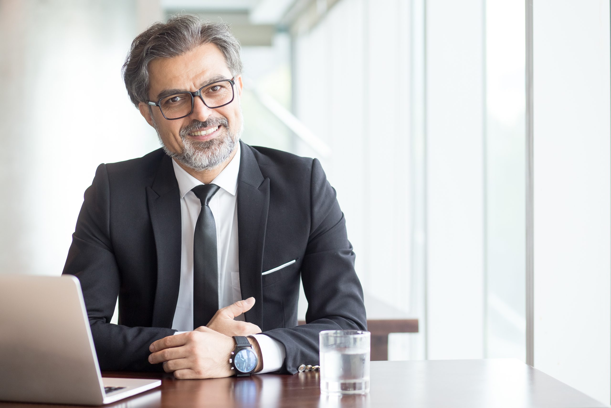 Cheerful businessman in eyeglasses working in office or coworking space. Portrait of confident male executive with gray hair at table. Successful business concept